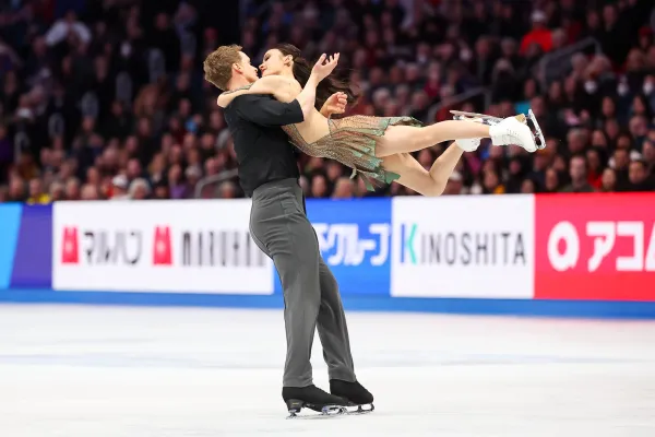 Male figure skater holds up female figure skater while performing on ice with a large crowd watching