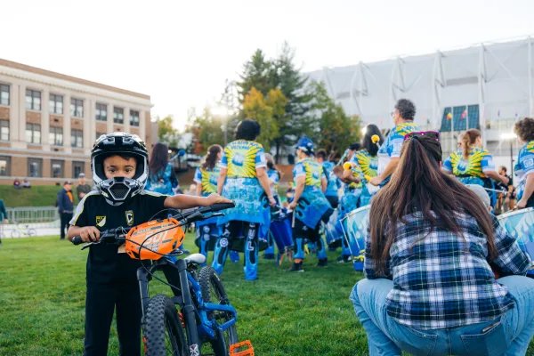 Young child looking at camera holds small bike and wears full Mountain Bike helmet in front of drum line performers
