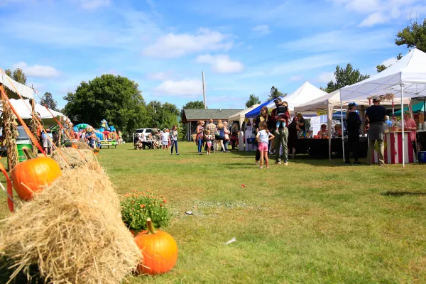 People browse at vendor tents in the background at outdoor event with pumpkin and hay bales in foreground