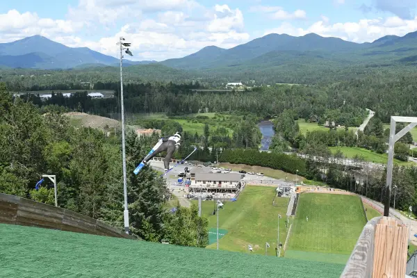 Ski jumper seen flying through the area above turf jump track in summer with mountains and venue in the distance