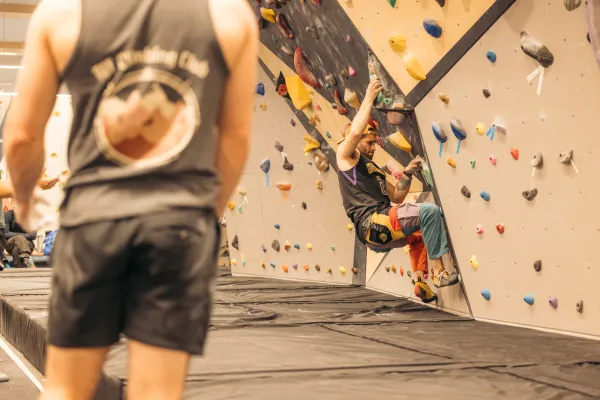 Climber on indoor bouldering wall with person looking on in foreground