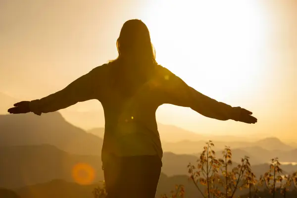 Stock image of woman's silhouette with outstretched arms at golden sunrise with faded mountains in distance