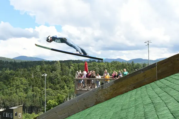 Ski jumper takes off of turf surface in summer while observation deck full of people looks on