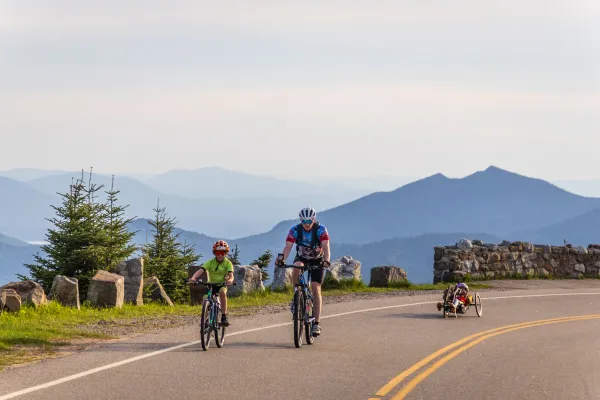 A cyclist and a child on a bicycle ride along a road with scenic views, followed by another person in a recumbent handbike.