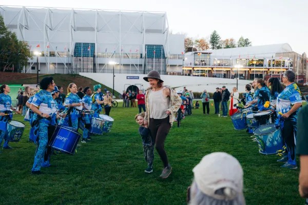 Woman holds child's hand as they run through the middle aisle of a drum line on the Olympic Oval in Lake Placid