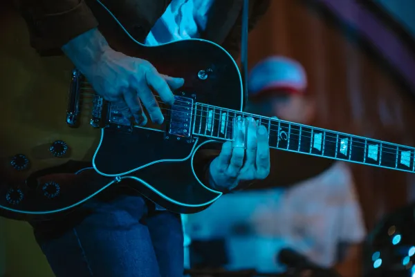 Close-up of person playing electric guitar on dark stage with drummer in background