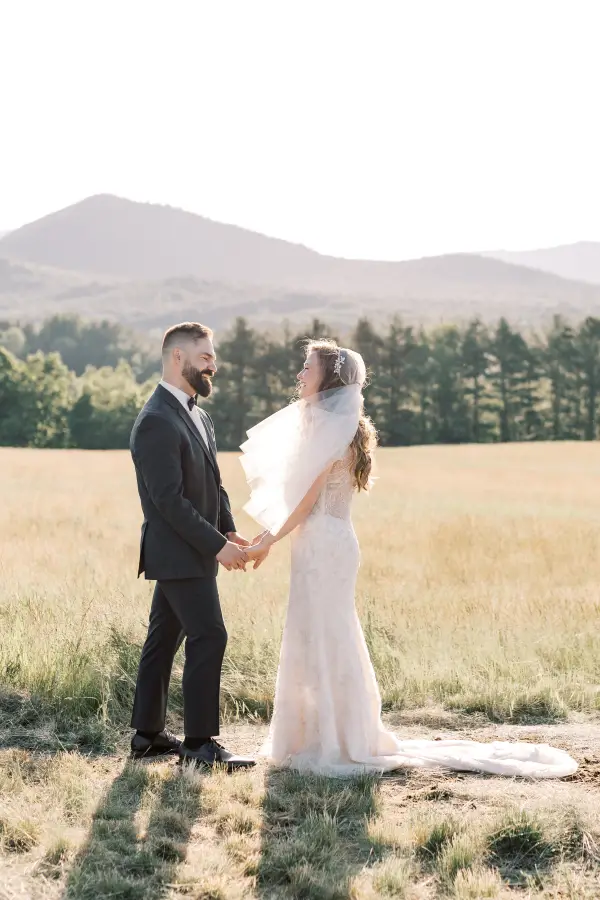 Bride and groom smile at each other in a wheaty field with veil blowing in the breeze