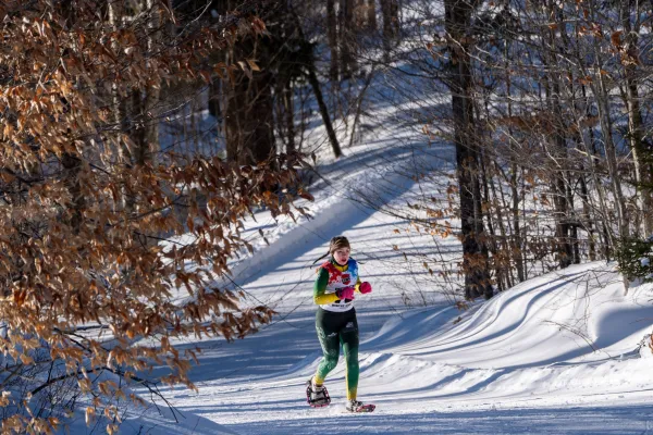 A young woman snowshoeing on a snowy trail, wearing vibrant athletic gear, under a clear blue sky with trees in the background.