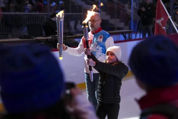 A young girl in a white beanie holds a flaming torch, assisted by an older man in a blue and red jacket. They stand indoors on an ice rink, surrounded by onlookers. The scene feels ceremonial and celebratory.