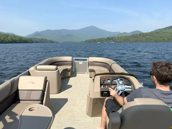 Man drives a pontoon boat on a lake towards a mountain in the distance