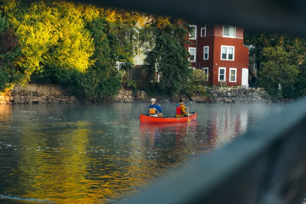 Man and woman paddle a red canoe down a river with a red house and trees on the bank