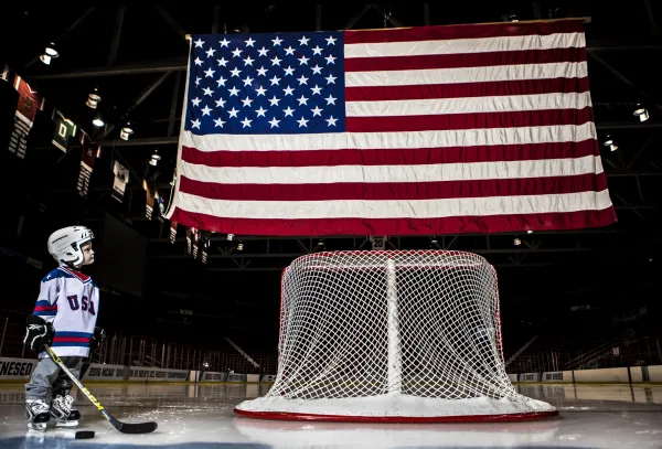 Young boy in hockey gear stands next to hockey goal faced away with large American flag flying above it in hockey rink on ice