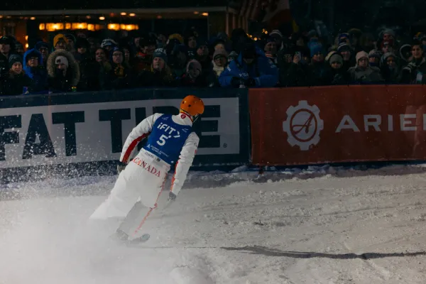 Freestyle aerialist skier skis into finish while a crowd looks on over a barrier at nighttime event