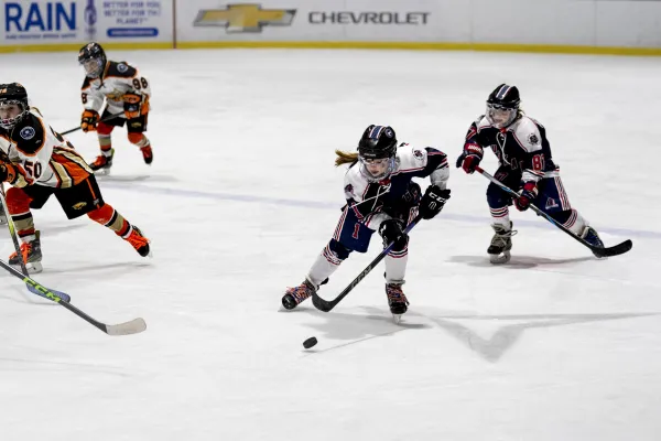 Exciting U10 girls' ice hockey match: a focused player in a white and navy uniform skates with the puck, pursued by teammates and opponents.