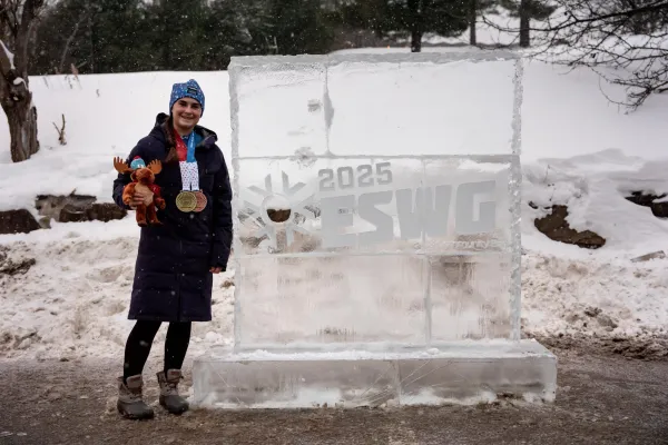 A smiling athlete stands proudly by a large ice sculpture for the 2025 ESWG, holding medals and a plush moose toy.