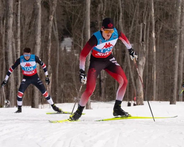 The image shows two cross-country skiers racing on a snowy trail. 