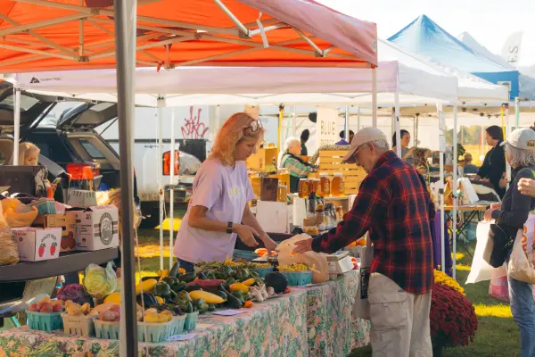 Older man purchases fresh produce from woman under red pop-up tent at outdoor farmers market