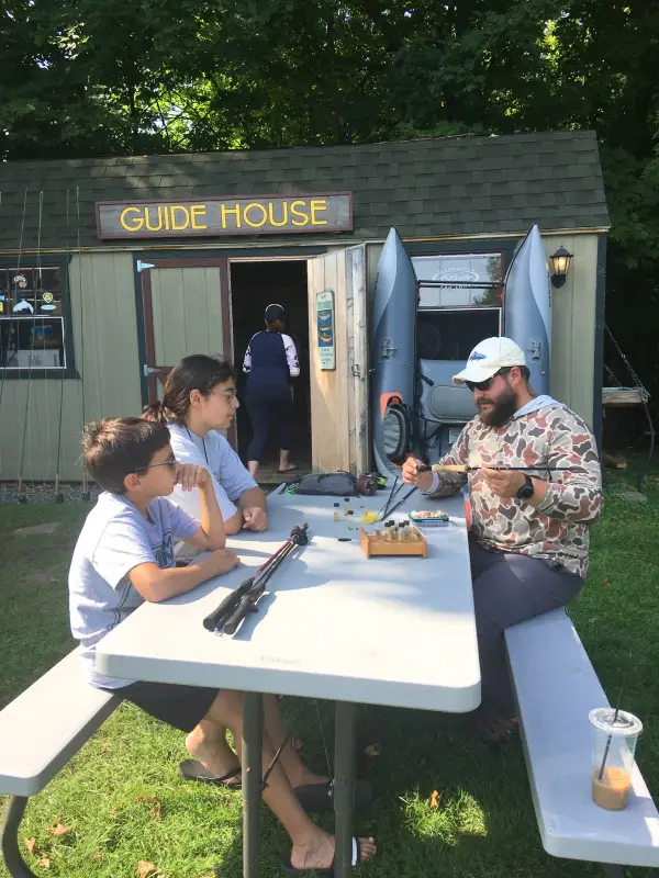 A family sits at a picnic table outside a guide house, preparing fishing gear.