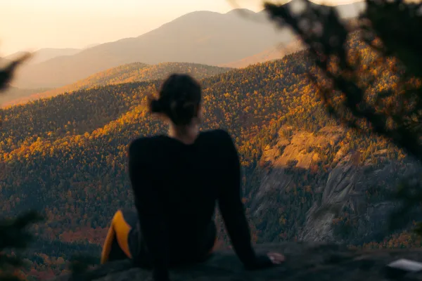 A person sitting with their back to the camera, looking out over a vast, layered landscape of mountains and forests colored in the gold and orange hues of autumn at sunset.