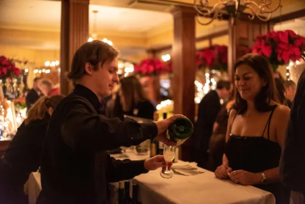 Bartender in dress clothes pours sparkling wine into a flute glass for a woman in a black dress on the other side of the bar at a busy party
