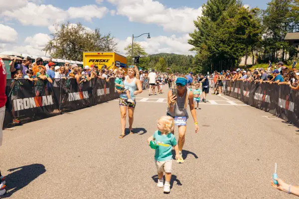 Woman walks behind child filming them and second woman carries toddler on Ironkids race course while spectators cheer