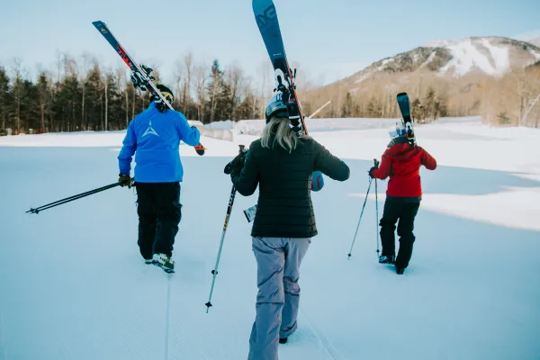 Three skiers in blue, black, and red jackets walk across a groomed trail carrying their skis over their shoulders.