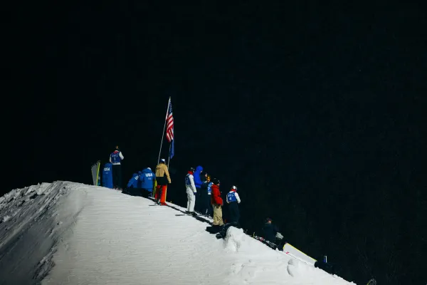 Group of people and skiers stand atop a snowy hill at night, illuminated with artificial light, looking outward into the dark empty space in front of them