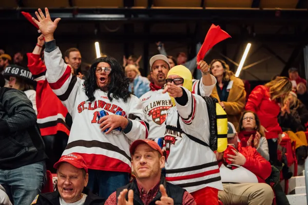 St Lawrence University hockey fans are seen standing in seats during game wearing jerseys and other costume items, cheering enthusiastically