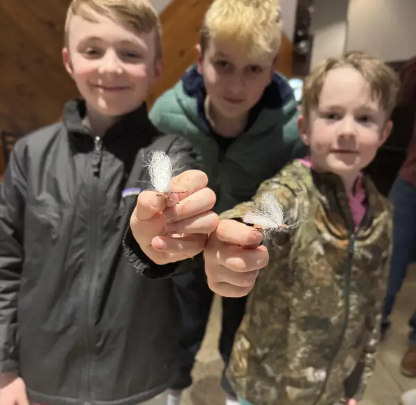 Two kids holding fly fishing flies up to camera while third child looks over their shoulders
