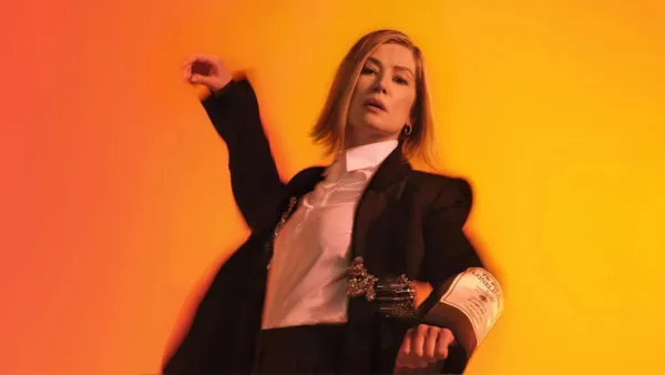 Rosemund Pike in a black and white suit dancing in front of yellow/orange background