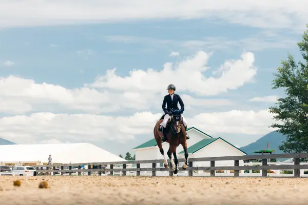 Equestrian competitor rides horse on dirt ring with white and green roofed building in background