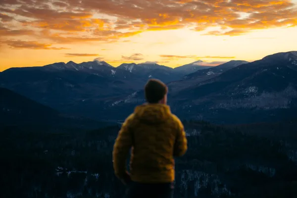 A person in a yellow jacket stands with their back to the camera, looking out over a mountain range in Lake Placid.
