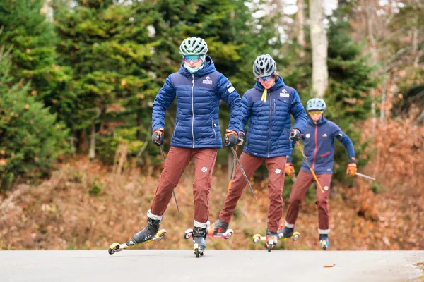 Three teenage student athletes practice cross-country skiing on specially equipped skate skis on a forested road.