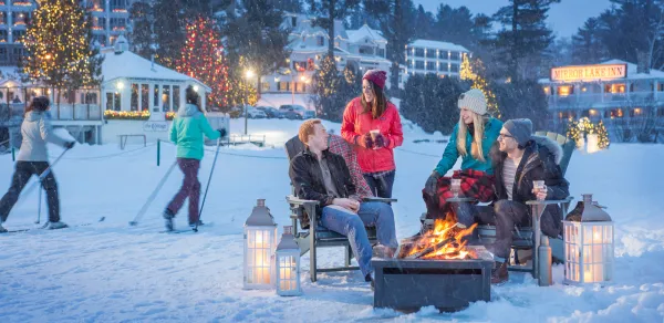 Happy people sit and chat around a fire pit with the glow of christmas lights dot the background and a skiier slides by.