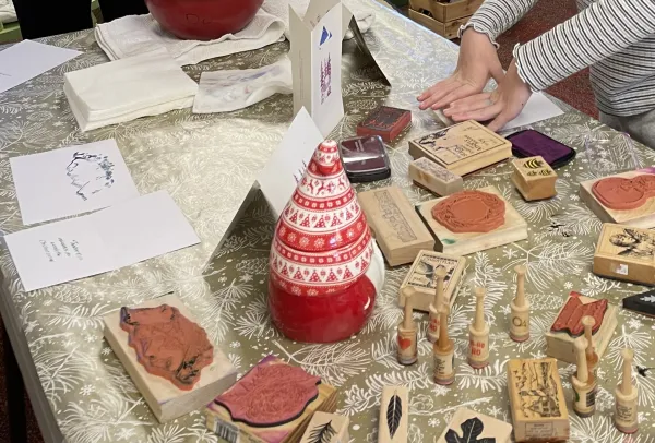 Table covered in various stamps, paper, a Santa-shaped ceramic decoration, and small hands pressing a stamp down on paper