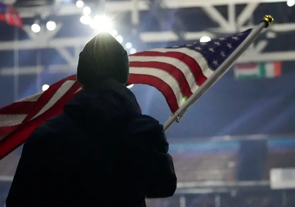 Person stands facing away from camera in a well-lit ice rink holding an American Flag on a pole
