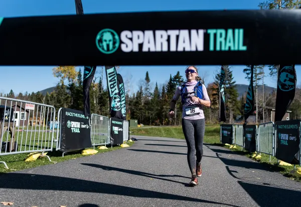 Woman wearing running vest smiles as she runs beneath a Spartan Trail arch over a paved path past event fencing