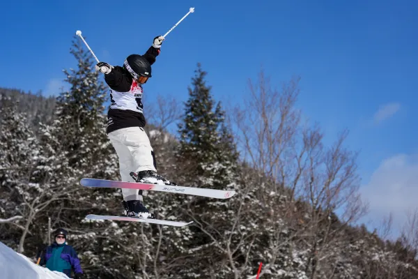 A freestyle mogul skier performs a jump with poles extended against a clear blue sky and snow-dusted evergreens.