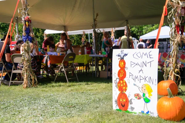 People sit under an outdoor tent with children with a sign out front that says "come paint a pumpkin"