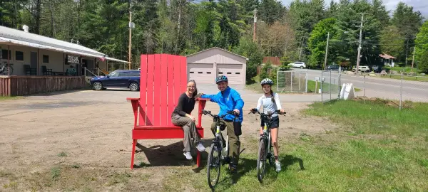 Three smiling people surround the giant red adirondack style chair out front of Bike Lake Placid