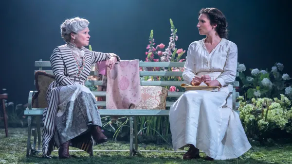 Two actresses sit next to each other on a bench looking at one another with a staged garden behind them. 