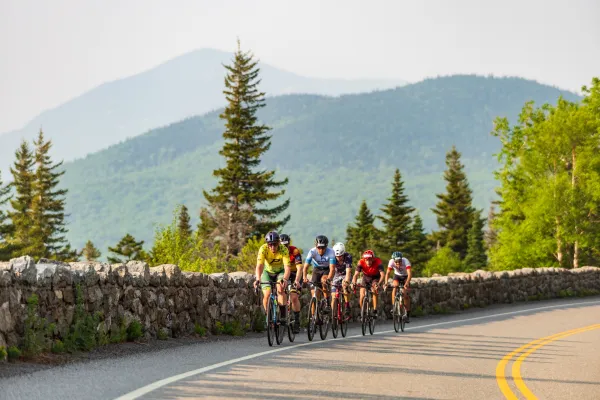 A group of cyclists rides in a tight pack along a winding mountain road bordered by a stone wall.
