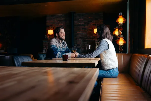 Couple sits across a wooden table with glasses of wine in an aesthetically dark restaurant with warm lamps and natural light