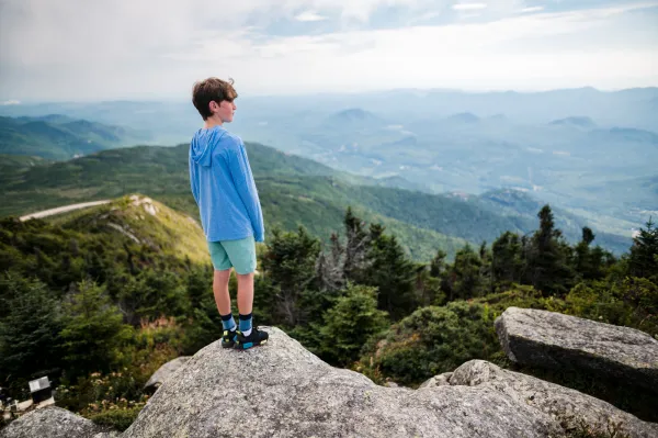A boy stands on top of a mountain overlooking the wilderness.