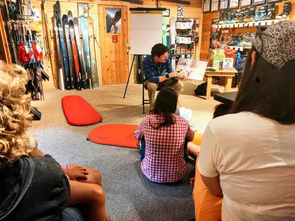 A man reads a children's book to a group of children sitting on the floor.  Ski equipment is visible in the background.