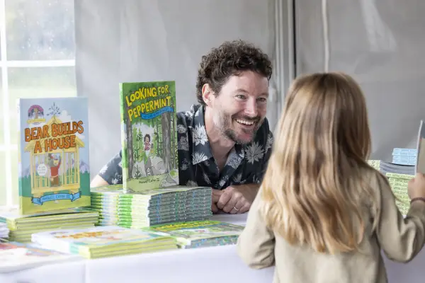 Middle aged white male author sits behind book display on table smiling at child standing across from him