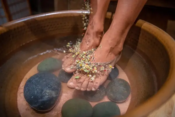 Feet immersed in a tub of steaming water, topped with herbs and some river stones within