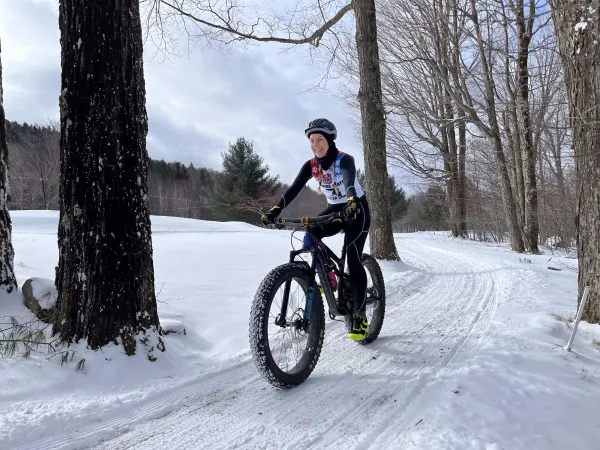 Person biking with a big smile on a snowy trail through a forest, wearing winter gear and helmet. The scene conveys excitement and adventure.