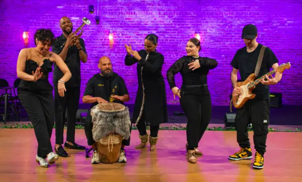 A band comprised of dancers and musicians performs on stage.  A woman tap dances in the foreground.  Other performers include a trumpet player, a drummer, two female dancers, and a guitarist.  Purple lighting illuminates a brick wall in the background.