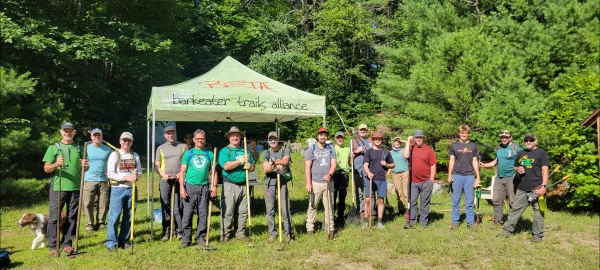 A group of volunteers stand in front of a Barkeater Trails Alliance tent, holding trail maintenance tools. 